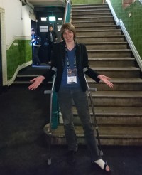 Photo of a woman on crutches standing at the bottom of a flight of stairs, shrugging her shoulders