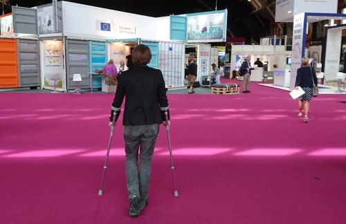 Photo of a woman in a black jacket and grey trousers, standing with the aid of crutches, preparing to walk into a large convention centre