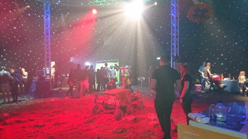 Photo of a rover prototype sitting on sand under red light inside a dome with a simulated starry sky